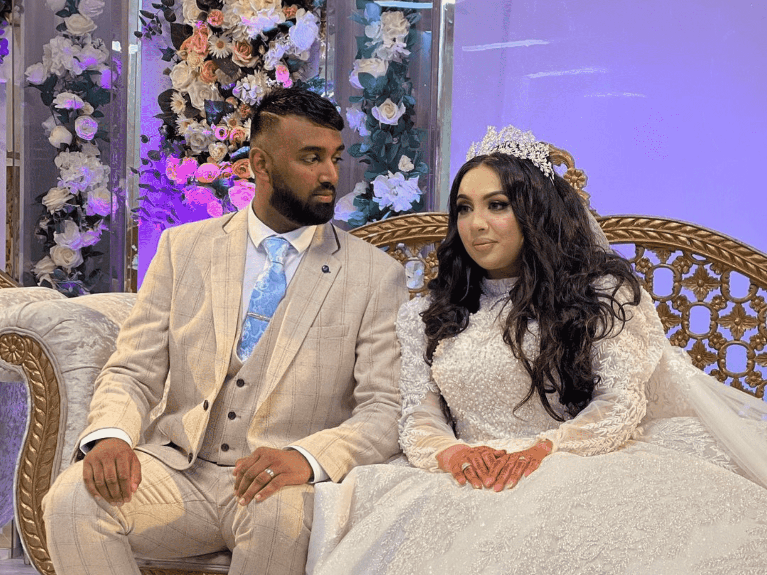 Bride and Groom sitting on a Gold Couch. Groom is wearing a nude colored suit with a blue tie. Bride is wearing a white dress.