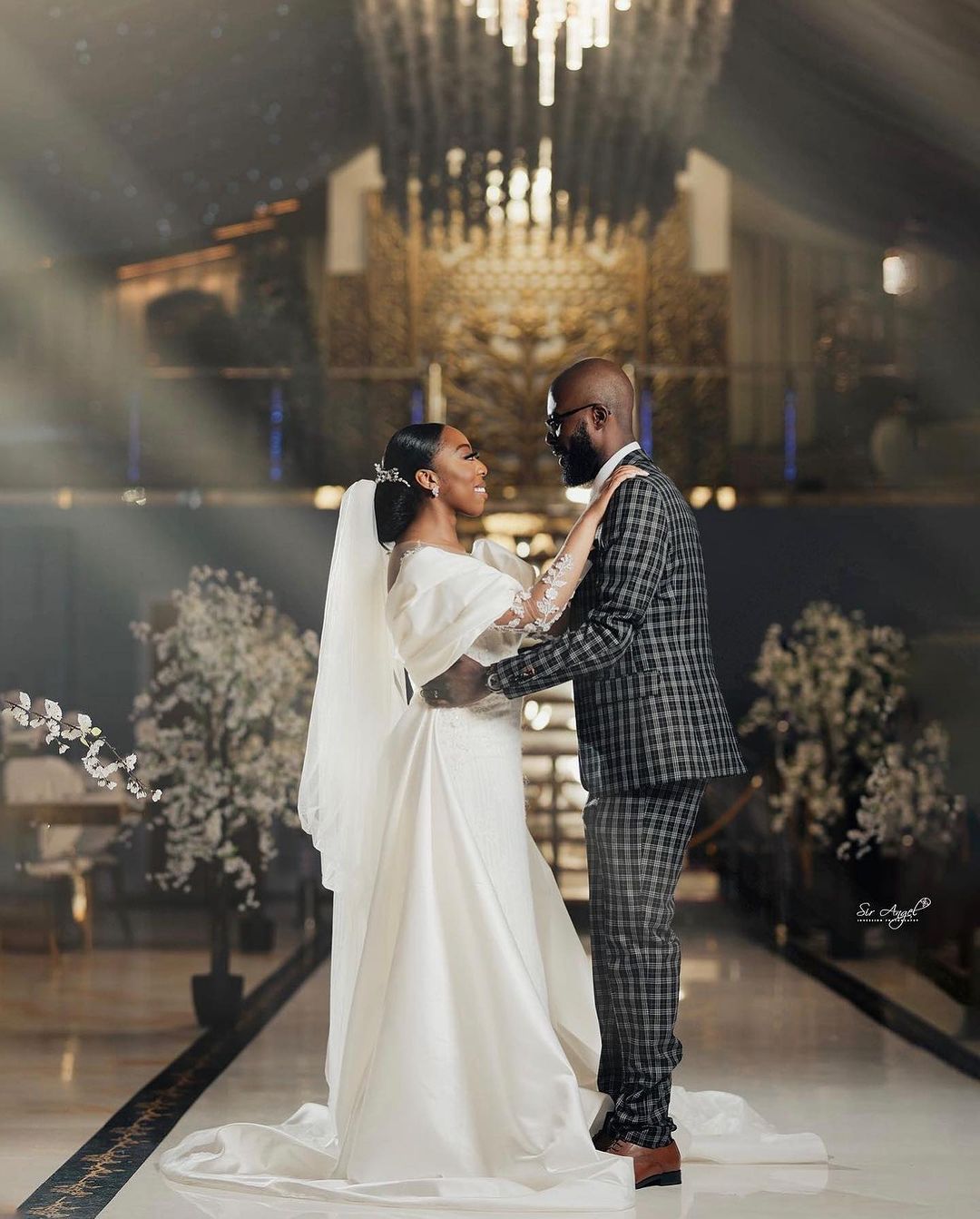 A bride and a groom dancing. Bride is wearing a white wedding dress. Groom is wearing a checkered suite