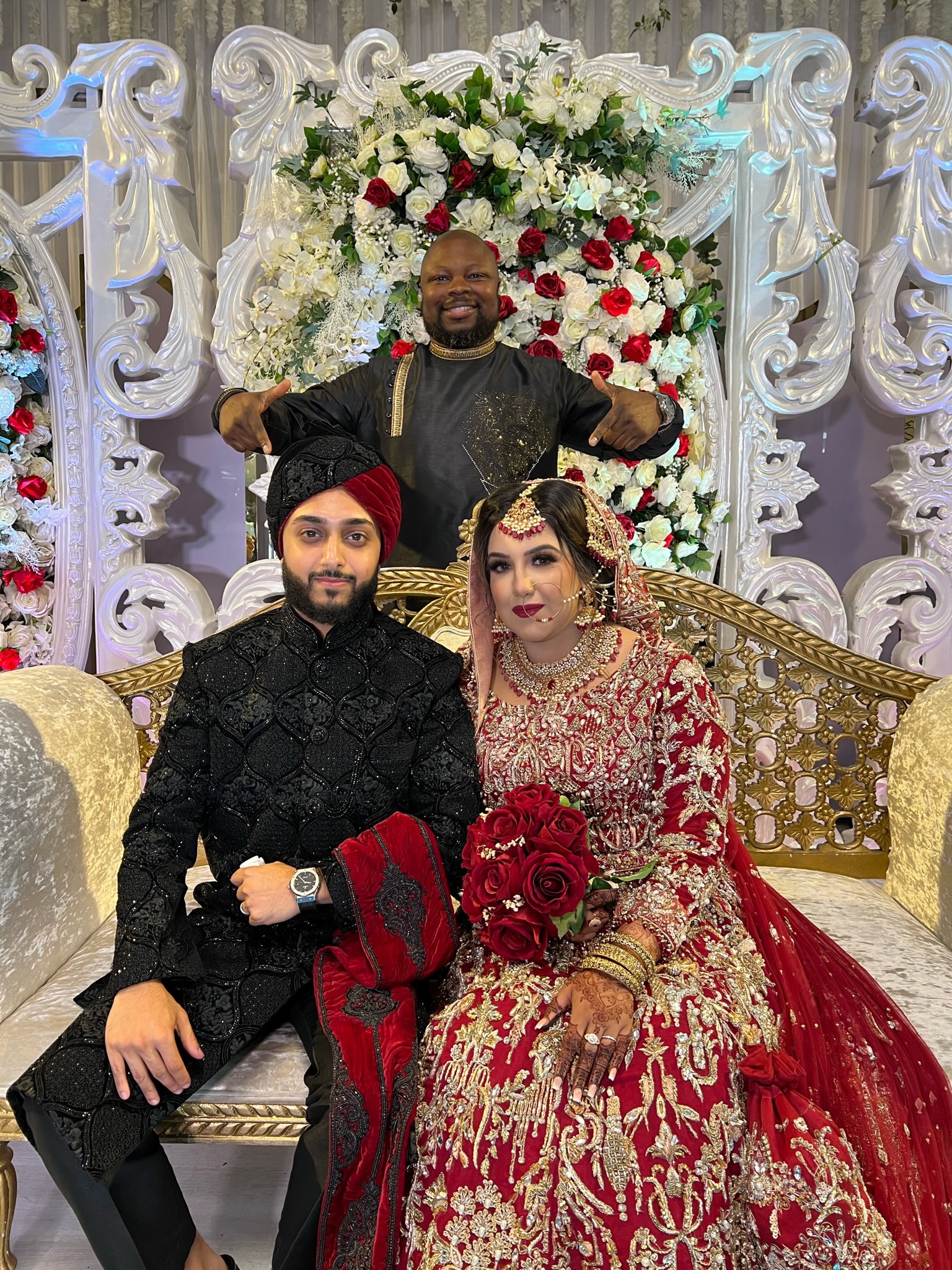 Bride and Groom sitting together on a Golden couch, Groom is wearing a black cultural outfit. Bride is wearing a red and gold outfit.