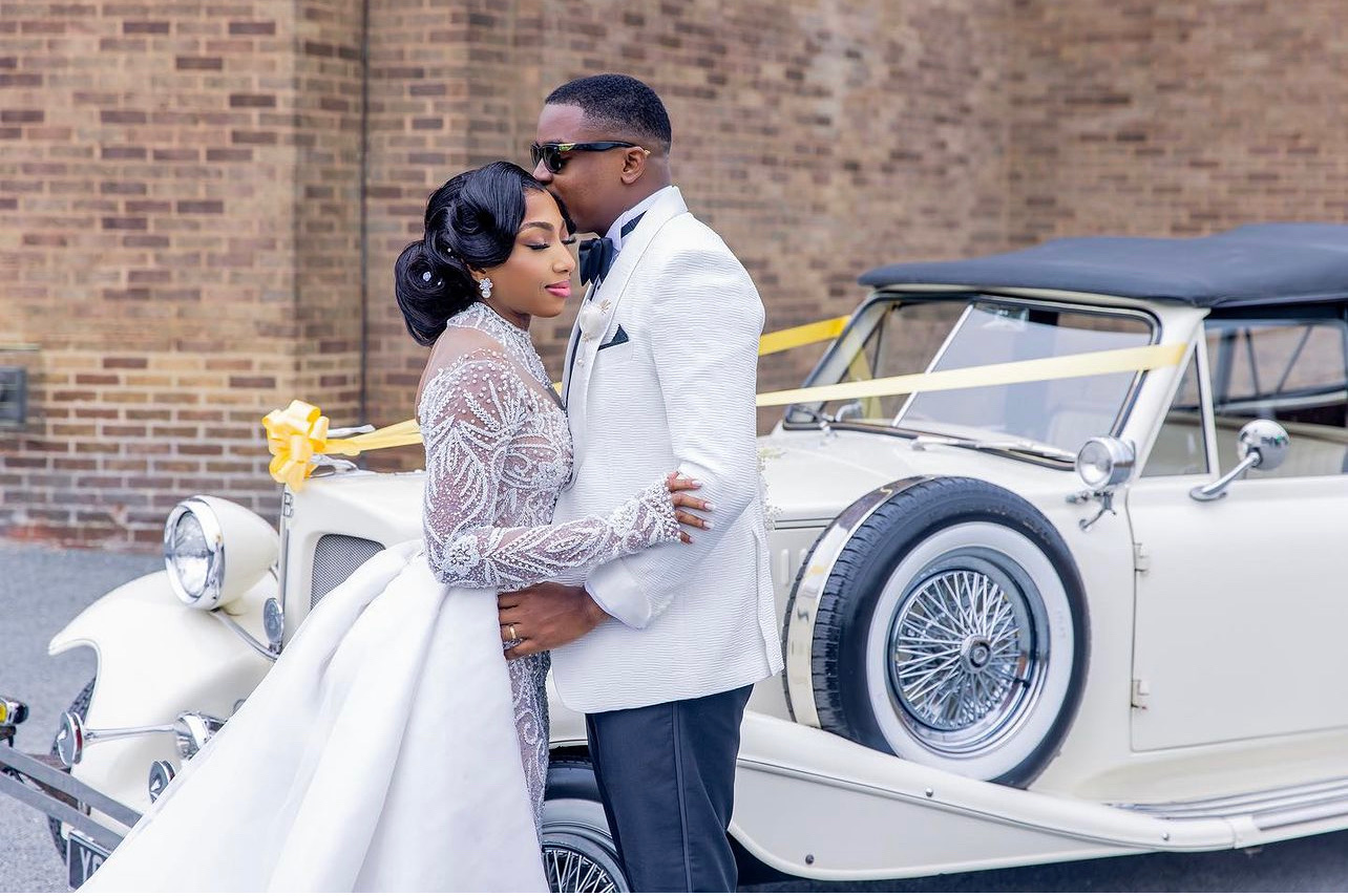 Bride and Groom holding each other close. They are both standing beside a white car. Bride is wearing a white beaded dress. Groom is wearing a white suit.